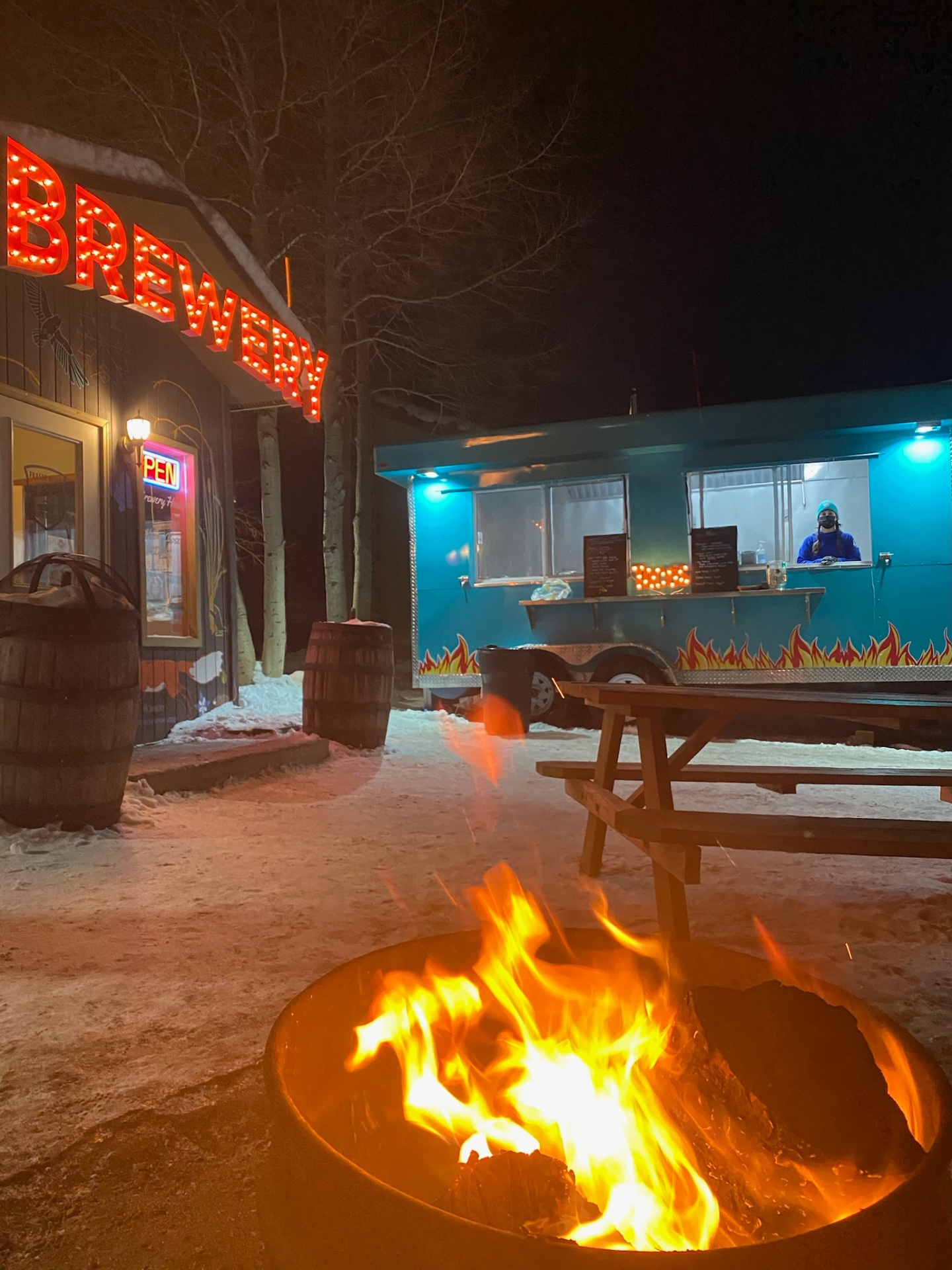 The food truck at night next to a brewery sign with a roaring fire pit in the foreground
